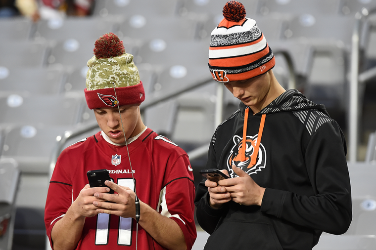 Two sports fans in team beanies and jerseys stand in stadium seating, looking down at smartphones.