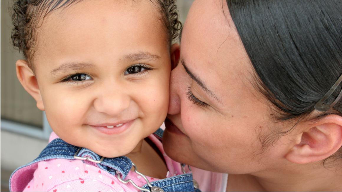 A mother kisses her toddler, who is looking at the camera