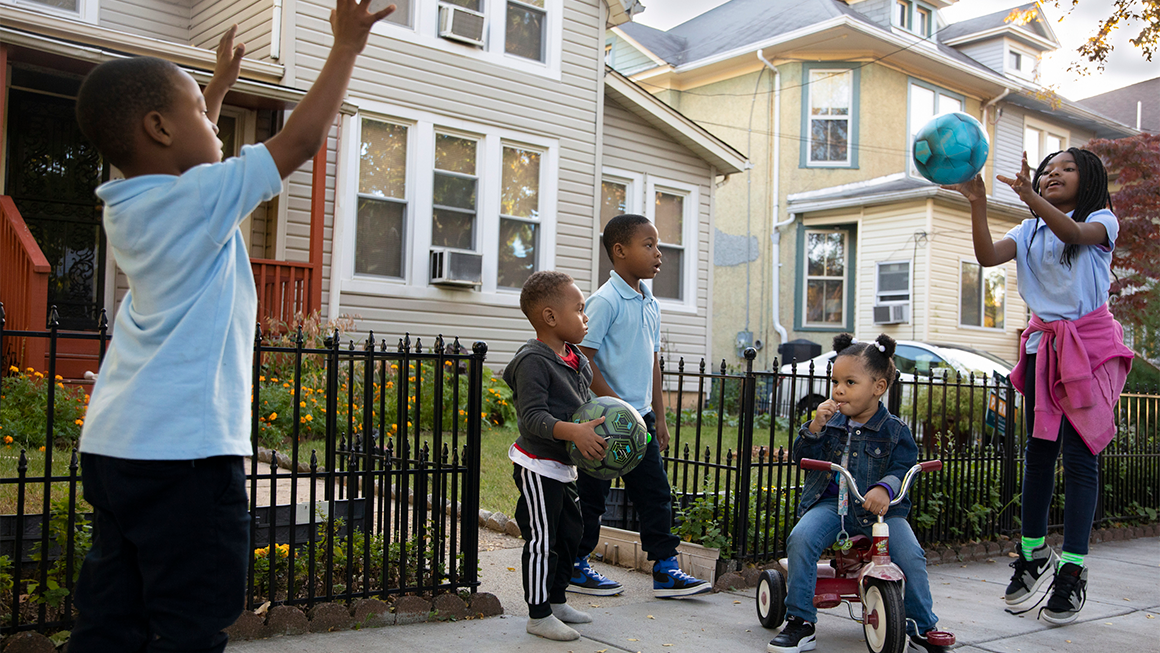 Children play ball on a sidewalk outside some homes on a sunny day
