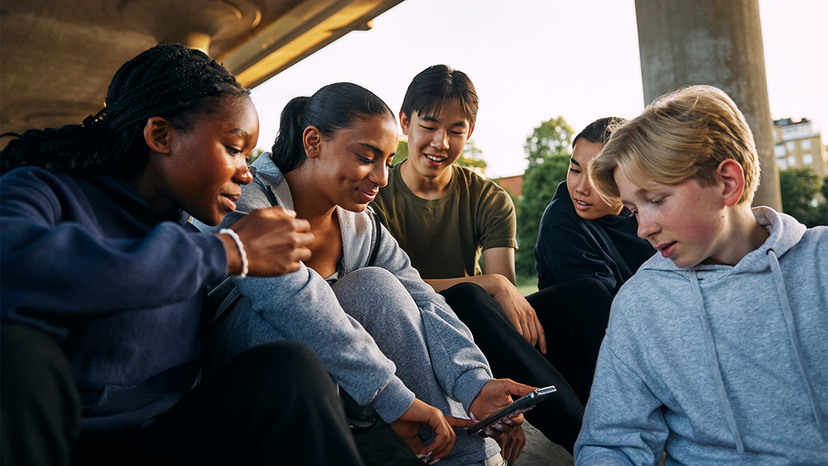 Five young people sit together outside, gathered closely around a smartphone.