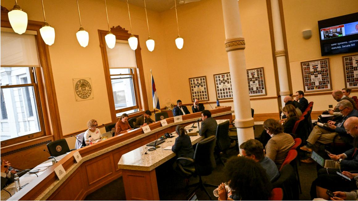People seated around a curved committee dais during a legislative hearing in a large government meeting room with high ceilings, tall windows, and a Colorado state flag.