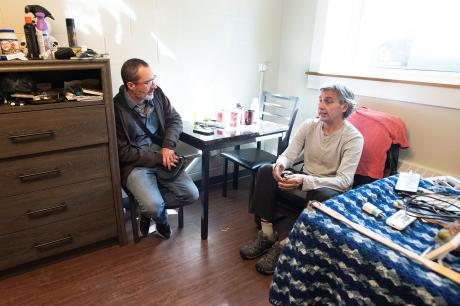 Two men sit in a room at a table beside a bed with personal objects on it