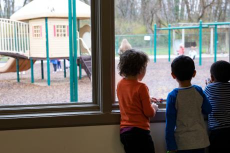 Three children stand at a school window looking outside at a playground