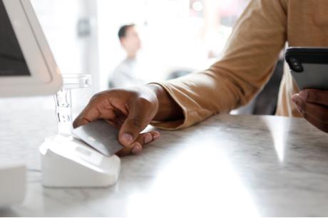Photo of someone tapping their credit card on a credit card reader as they look at their phone. 