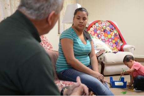 A man and young mother sit on a couch as her toddler son plays on the floor nearby
