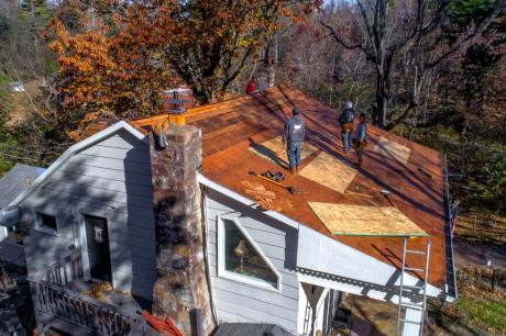 Photo of four construction workers redoing the roof of a small house in a wooded neighborhood.