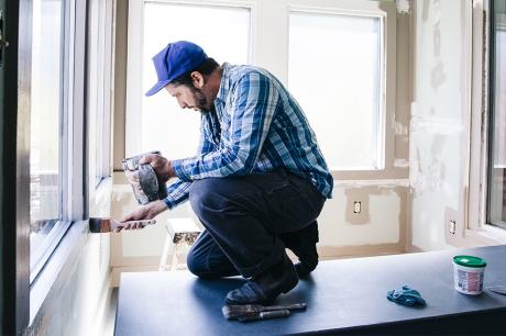 Photo of a man kneeling on a table and painting window trim.  