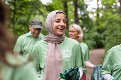 A smiling woman wearing a beige hijab and a green volunteer T-shirt stands outdoors in a wooded area, holding a trash bag, while other volunteers in matching shirts gather around her during a park cleanup.