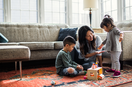 A mother and two toddler children play on the floor of a living room 