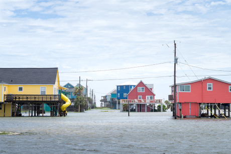 Floodwaters surround colorful stilt houses in a coastal neighborhood, with water covering streets beneath an overcast sky.