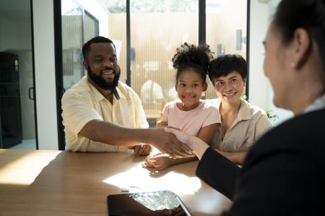 Person seated at a table shakes hands with a representative while two other people sit beside them in a bright office.
