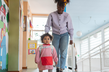A young child walking through the halls of a day care, followed by her mother.