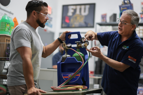 Two people in a workshop review HVAC refrigerant gauges connected to gas cylinders during hands-on technical training.