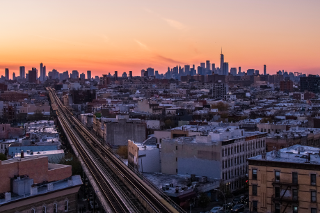Panoramic city skyline at sunset, with elevated train tracks leading through low-rise buildings toward a cluster of downtown skyscrapers under a pink-orange sky.