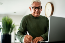 Image of an older man in a green shirt writing something down on a paper next to his laptop.