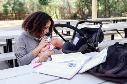 A young mom studies for a college exam outside at a picnic table while feeding her baby a bottle