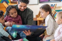 A daycare educator sits on the floor with a child on his lap and other toddlers scattered around him as he reads them a story. They are each dressed casually and are focused on the book.