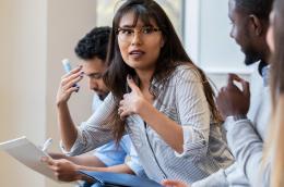 A woman with dark hair and glasses engages in a group discussion.