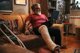 A man in a red shirt sitting on a leather sofa balances a laptop in his left hand, using a mouse with his right, and his left leg is in a cast, propped up on a stool next to a pair of crutches.