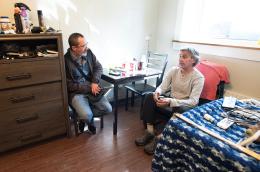 Two men sit in a room at a table beside a bed with personal objects on it