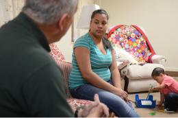 A man and young mother sit on a couch as her toddler son plays on the floor nearby