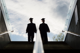 Two graduates in caps and gowns walk down a wide outdoor staircase, silhouetted against a bright sky, with glass railings and concrete walls on either side.