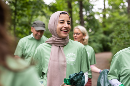 A smiling woman wearing a beige hijab and a green volunteer T-shirt stands outdoors in a wooded area, holding a trash bag, while other volunteers in matching shirts gather around her during a park cleanup.