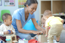 photo of daycare teacher sitting on the floor of a classroom with three sweet little toddlers, as they play together with toys