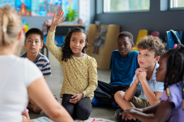 Photo of a young girl of color raising her hand as she and other students sit on a rug with their teacher. 