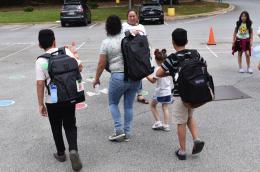 A family of adults and children wearing backpacks walks away in a parking lot 