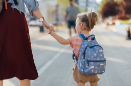 A young child wearing a small blue backpack holds an adult’s hand while walking down a sunlit city sidewalk