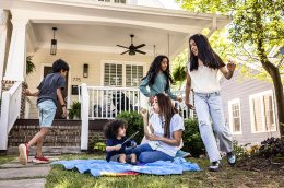 People gather outside a house while someone blows bubbles for a child on a sunny day.