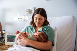 A young mother reclined in a hospital bed smiles down at her baby, as a person in the background adjusts medical equipment.
