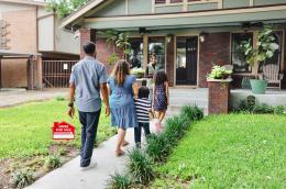 A family of four walks past a "Home for Sale" sign toward a smiling realtor and a red-brick home with an open front door.