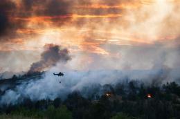 A helicopter carrying  a helicopter bucket of water is silhouetted against a sunset and plumes of smoke, with a wildfire burning a forest below.
