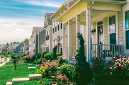 A quiet suburban street with modern two‑story houses, each with front porches, trimmed lawns, and colorful landscaped gardens. 