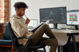 a young man working at a desktop computer at home, reading a book while analyzing financial charts on the screen