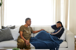 An older adult sits on a couch gently resting a hand on a younger person who is lying under a blue blanket