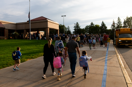 Families and children with backpacks walk toward a school entrance as a yellow school bus waits.