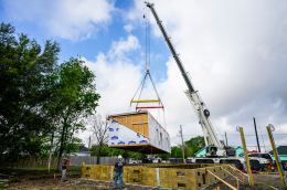 Workers guide a crane‑lifted modular home section onto its foundation at a construction site