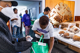 People collect food items at a community distribution table, with volunteers placing packaged items into a green bag; boxes and loaves of bread are visible nearby.