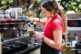 A young woman with long ombre hair, pretty orange earrings, and an apron tied over a red t-shirt pours a shot of espresso into a stonewear mug in a cafe with a whole wall of plants.
