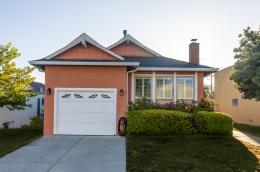 An outside view of a small orange house with a white garage door and well-kept grass lawn that features some shrubs, plants, and flowers.