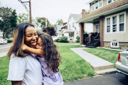  A happy little girl hugs her mom, who is smiling so hard her eyes are closed and her nose is crinkling, stand on the driveway in front of a two-story home.