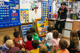 An educator stands at the front of a colorful classroom, reading to a group of young children seated on a carpet, surrounded by learning charts, books, and classroom displays.