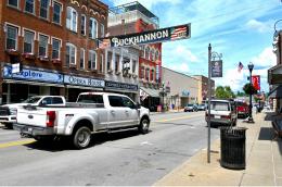 Downtown street in Buckhannon, West Virginia, with brick storefronts, parked vehicles, and a large “Buckhannon” banner hanging above the road.