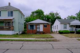 Small brick-and-siding house between two larger homes on a residential street, with a driveway, lawn, and overcast sky.
