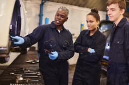  An woman in a long-sleeve navy jumpsuit and light blue latex gloves holds an oily automobile part in her left hand while gesturing to a part in the wheel well of a suspended white automobile as two students in jumpsuits and gloves look on attentively.