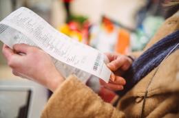  A close-up shot of a woman in a camel-colored coat and navy blue scarf holding a very long receipt, with the top half folded behind the bottom, to inspect the total amount due.