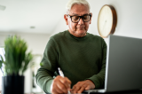 Image of an older man in a green shirt writing something down on a paper next to his laptop.